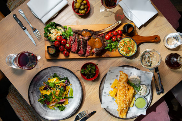 A table full of delicious food. One large steak platter in the centre with sauces and garnish. A fish and chips and a pasta dish. The table is set and there are glasses of wine. 