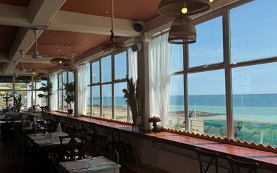an inside of a cafe with a sea view. outside the sun is shining and large windows overlook it. A mix of seating and tables. Saltdean Cafe and Restaurants