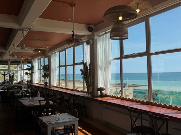 an inside of a cafe with a sea view. outside the sun is shining and large windows overlook it. A mix of seating and tables