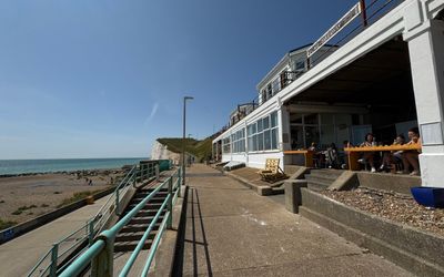 the exterior of a seaside cafe. The buiilding is white and carved into the cliff. In the background you see a white sea cliff and a blue sea.