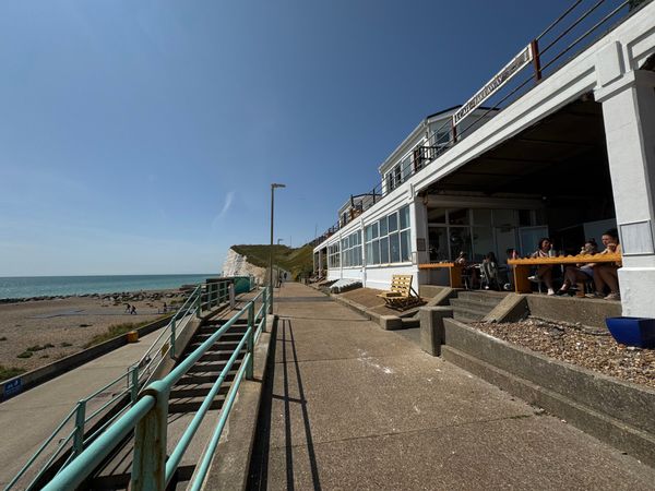 the exterior of a seaside cafe. The buiilding is white and carved into the cliff. In the background you see a white sea cliff and a blue sea.