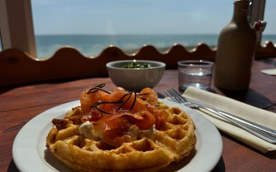 a brunch item which is a waffles topped with smoked salmon and cream cheese. It sits on a table at a window that overlooks the sea.