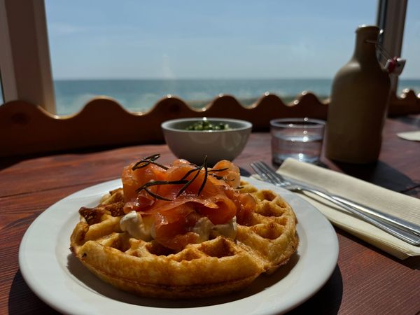 a brunch item which is a waffles topped with smoked salmon and cream cheese. It sits on a table at a window that overlooks the sea.