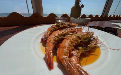a white plate sitting on a table is filled with king prawns. Outside the window the sky is blue and the sun is shining in making the prawns look pinks and gorgeous.