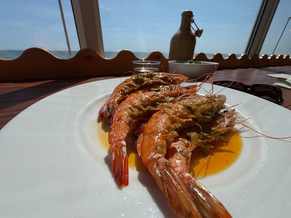 a white plate sitting on a table is filled with king prawns. Outside the window the sky is blue and the sun is shining in making the prawns look pinks and gorgeous. 