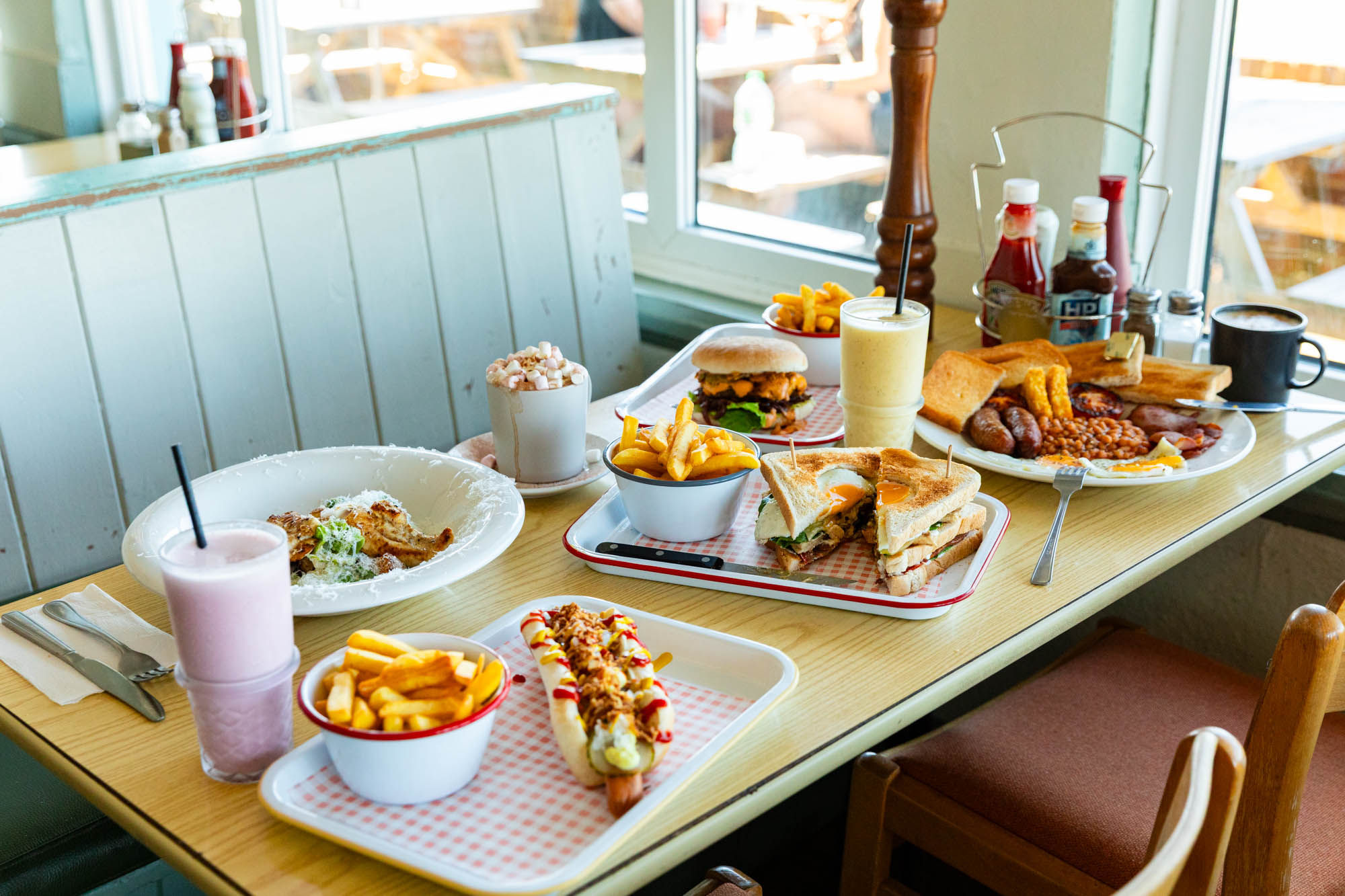 window table laid out with multiple dishes including toasts with chips, burger with chops, few drinks and sauces