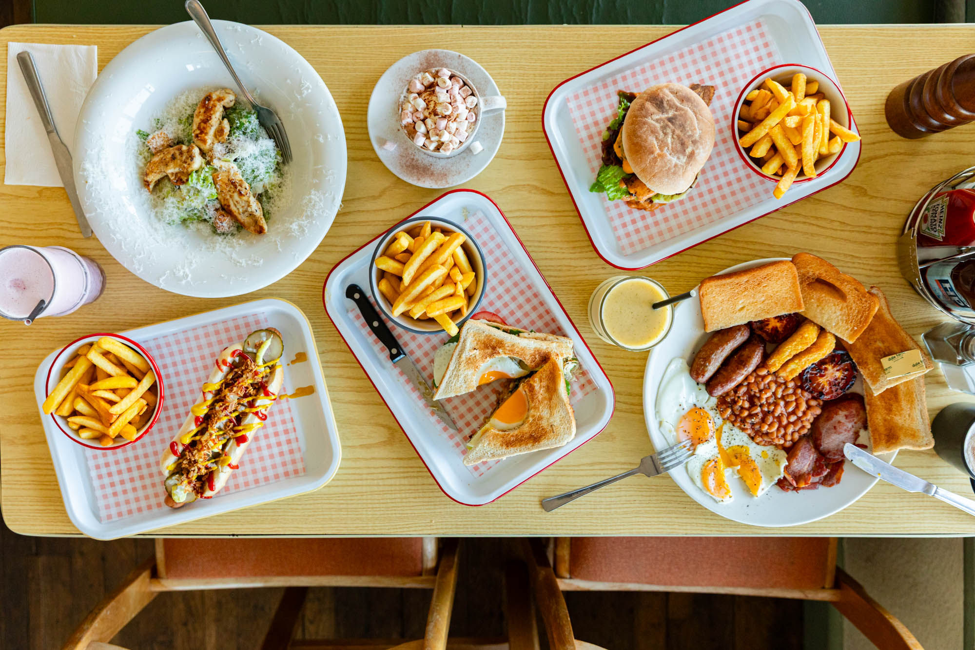 over head shot of the wooden table at Roedean Cafe populated with various dishes including full english breakfast, burgers and chips, toasts and chips, coffee and smoothies