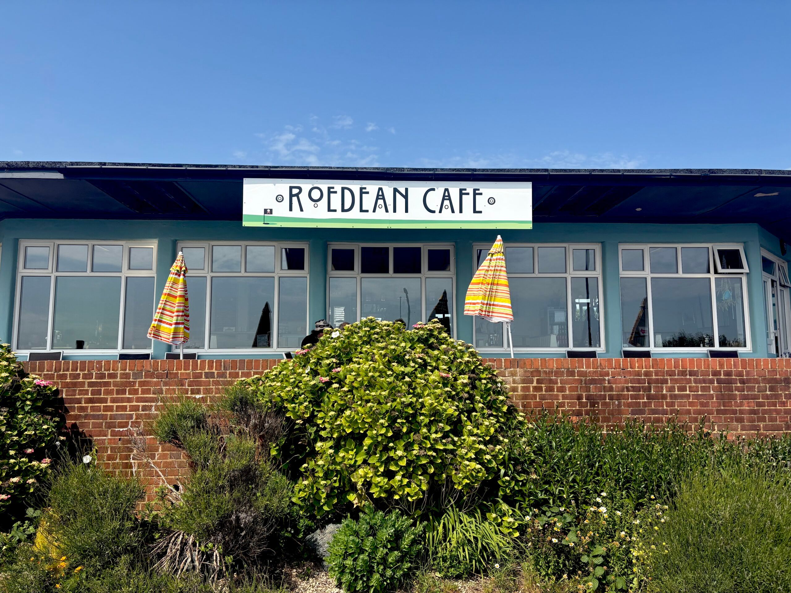 An exterior photo of a cafe against a blue sky. In the foreground is shrubbery and table umbrellas. The cafe has big windows and a sign in art deco lettering that says Roedean Cafe