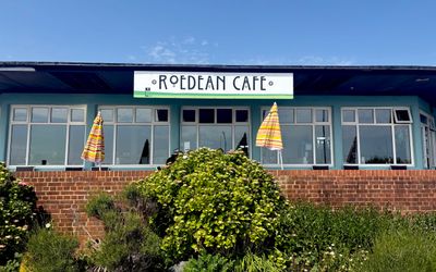 An exterior photo of a cafe against a blue sky. In the foreground is shrubbery and table umbrellas. The cafe has big windows and a sign in art deco lettering that says Roedean Cafe