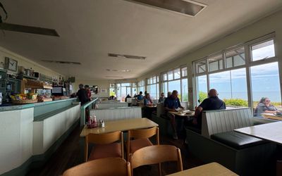 The inside of a cafe. Tables with chairs fill the fram. The right hand wall are windows looking out to sea. The left hand is the cafe counter.