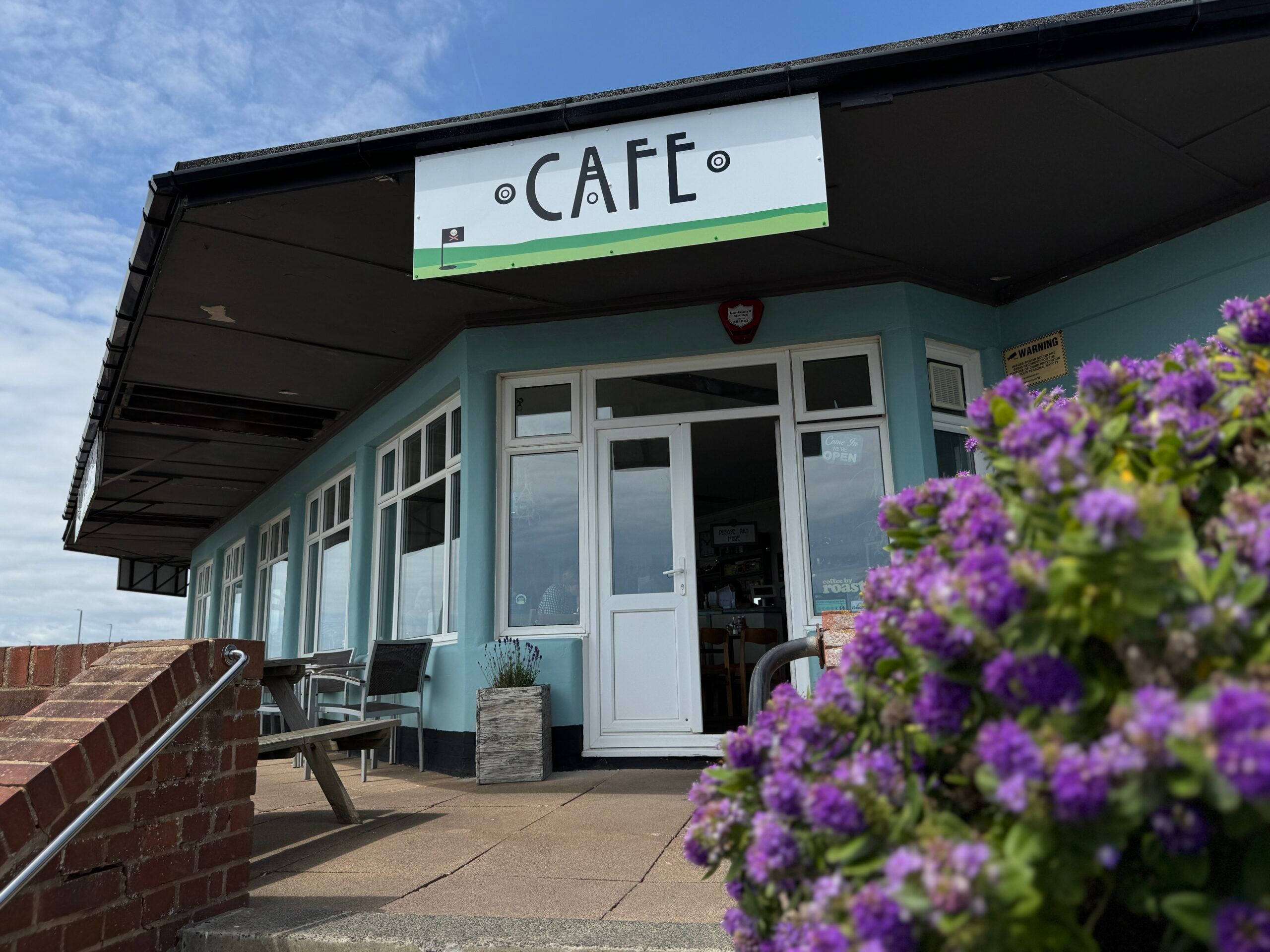 a building with a sign reading cafe is mainly in shot. It's a blue sky with a flowering bush in the foreground. 