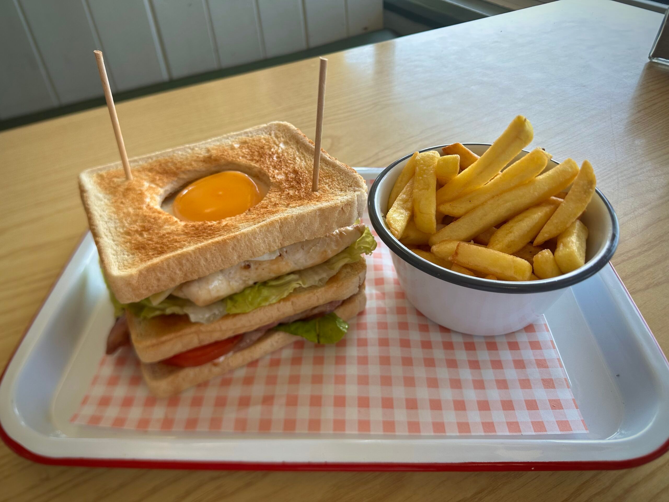 a club sandwich made with white bread layered with chicken and bacon topped with an egg sits on a tray. Next to it sits a bowl of french fries in a white bowl.