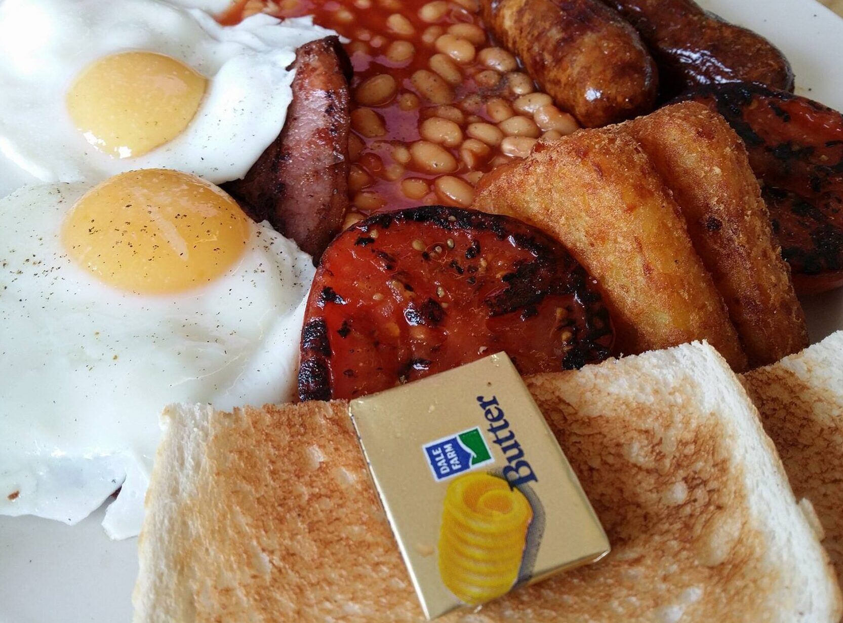 a close up picture of breakfast iterms. eggs, hash browns, beands, toast, sausages, tomatoes and a mini packet of butter.