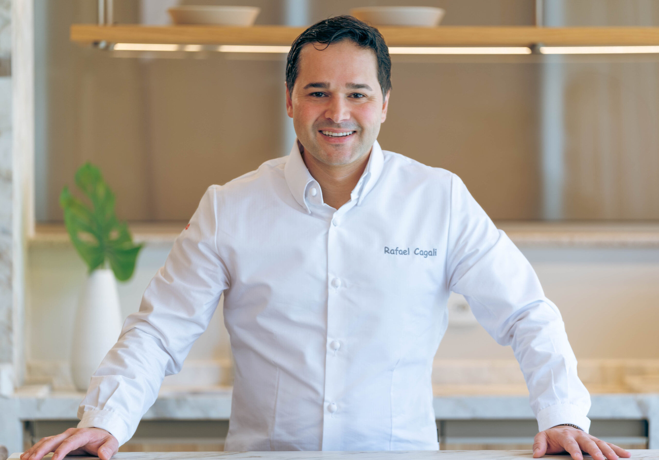 RAFAEL CAGALI - Marè restaurant in Hove, 1 Michelin Star in Brighton. Awarded February 9th 2026. Rafael dressed in chefs whites, stood in front of a kitchen counter with a plant in the background. He has his name embossed in his chef whites