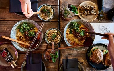 A wooden table is covered with asian style cuisine. Bowls of noodles and rice. Skewers and other delicious food is being scooped out for this picture. It is rustic in feel and we know that all the food is plant based.