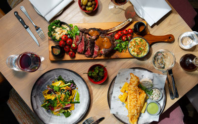 a table scene. Four dinner settings laid on a square table. One dish is fish and chips. a platter of steak and sauces sits in the middle. It all looks delicious.