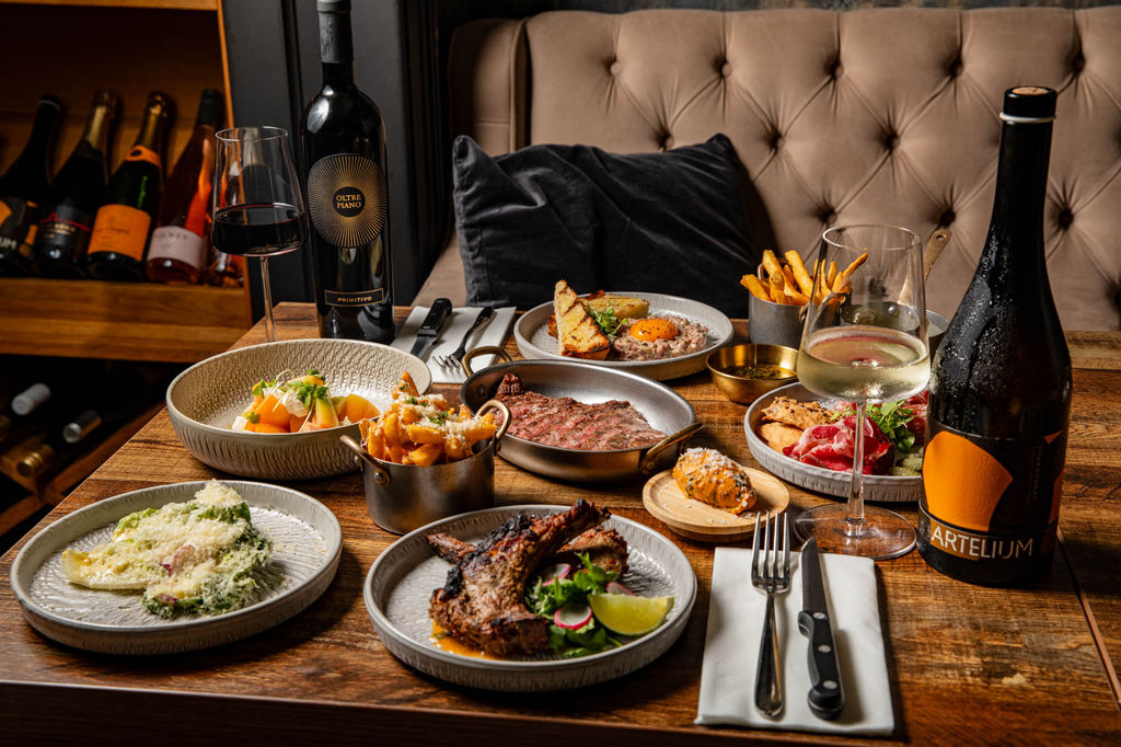A feast of food on a wooden table with bottles of wine and a black plush cushion in the background.