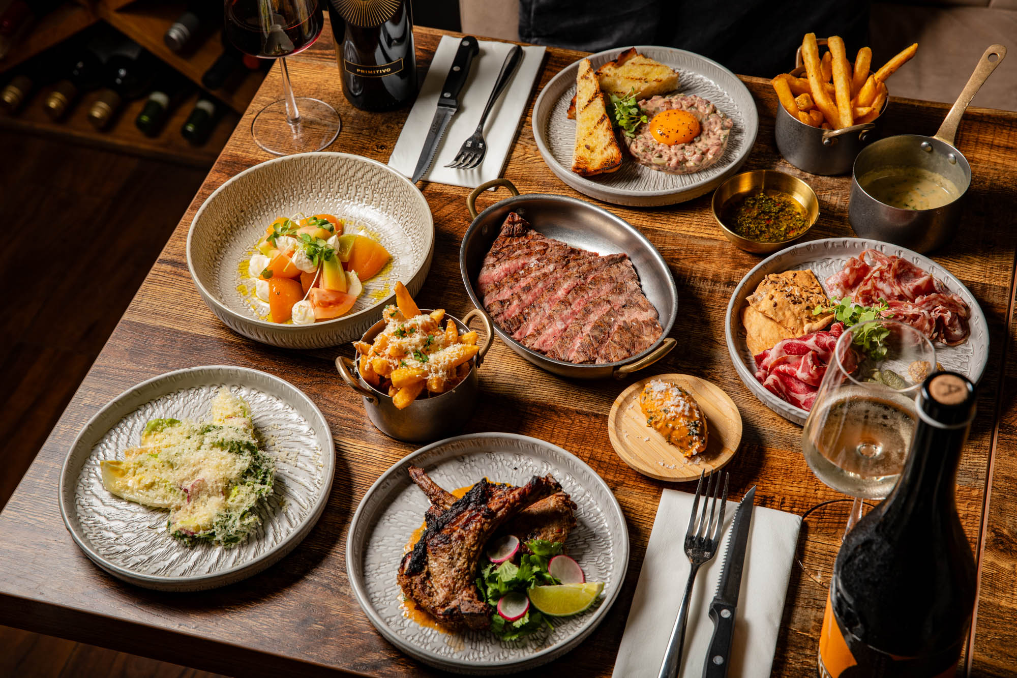 wooden table at Carne Hove laid out with six delicious dishes with sides, including steak, fries, charcuterie, tartare and more.