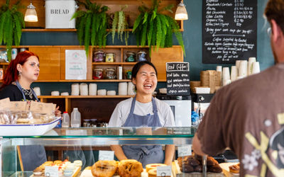 A counter of a bakery with a friendly server smiling at a customer. There is a glass conter that has lots of sweet treats in it. It looks like a friendly and welcoming place.