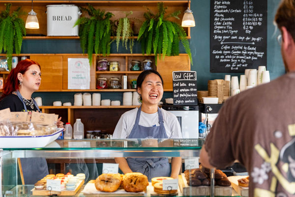A counter of a bakery with a friendly server smiling at a customer. There is a glass conter that has lots of sweet treats in it. It looks like a friendly and welcoming place.