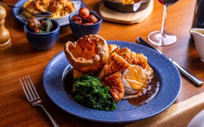 A Sunday Roast at Coppa Club Brighton A blue plate with a yorkshire puddinge, greens, carrots and meat with gravy. In the background on the table there are sides in small dishes and a glass of red wine.