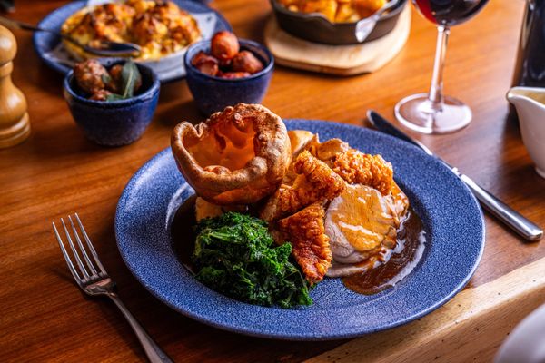 Sunday roast at Coppa Club Brighton. A blue plate with a yorkshire puddinge, greens, carrots and meat with gravy. In the background on the table there are sides in small dishes and a glass of red wine.