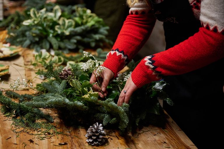 A person in a red jumper making a wreath with pine cones and foliage.