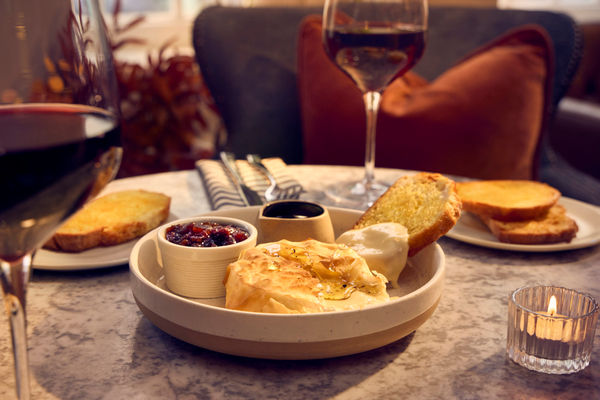 a dining table with a sharing plate with a baked camembert, some dips and sourdough toast. there are two glasses of red wine on the table too