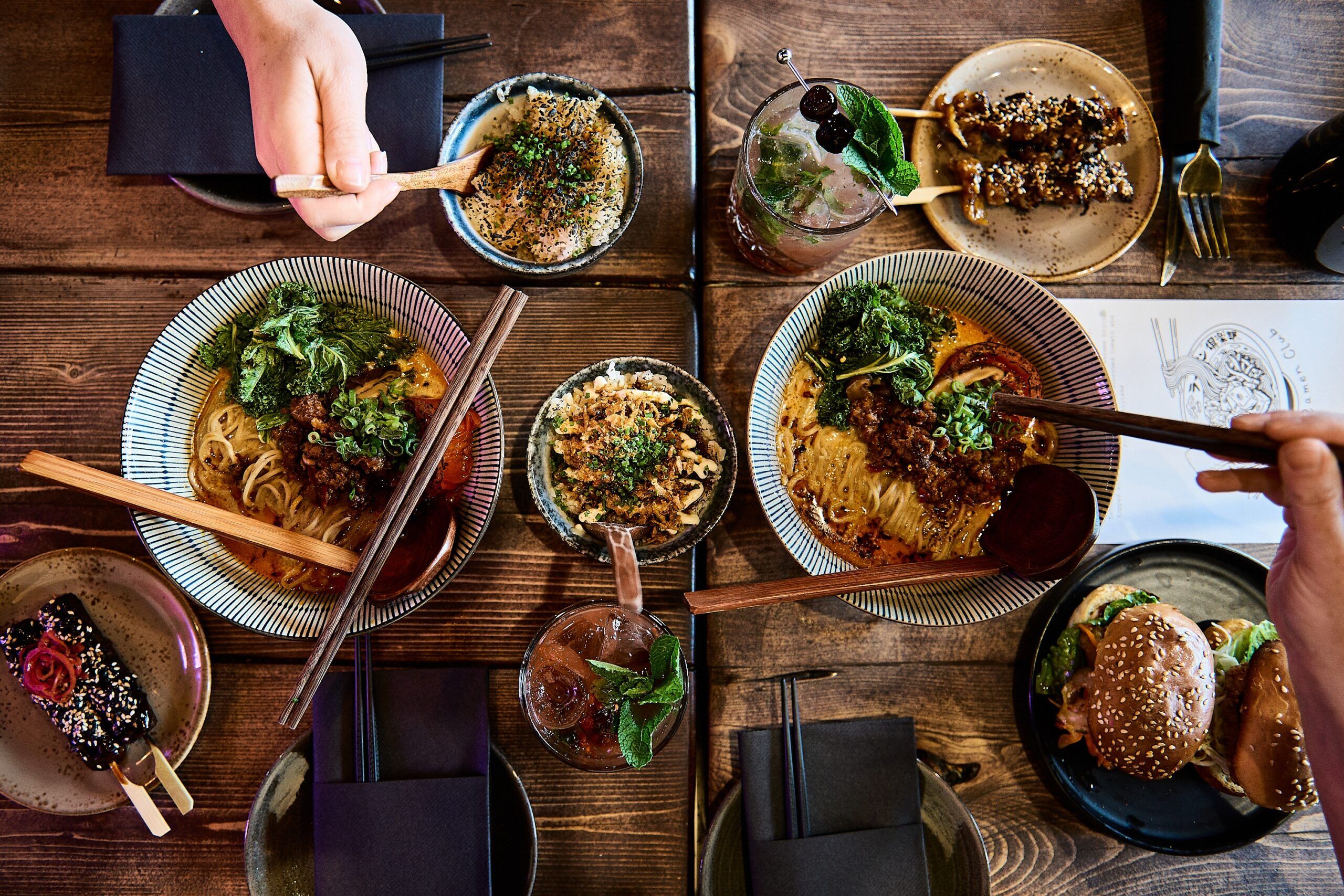 Overhead shot of a wooden dining table laid with two ramen bowls topped with greens and minced meat, two small rice bowls, skewers and a dessert stick, sesame-seed burgers on a plate, two cocktails, chopsticks and hands reaching in to eat