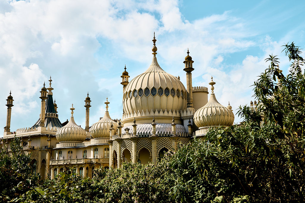 Brighton Pavilion against a cloudy blue sky with the sun shining on the golden building and leaves framing the pavilion.