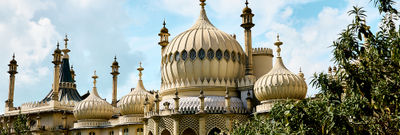 Brighton Pavilion against a cloudy blue sky with the sun shining on the golden building and leaves framing the pavilion.