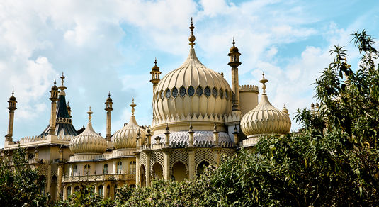 Brighton Pavilion against a cloudy blue sky with the sun shining on the golden building and leaves framing the pavilion.