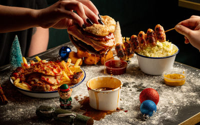 Peoples hands with long back painted nails holding a burger that's been dipped into a sauce and is dripping with a collection of other festive dishes and a few Christmas baubles on the table.