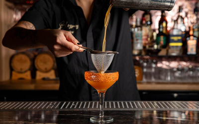 An orange cocktail being strained into a glass with orange coloured salt around the rim.