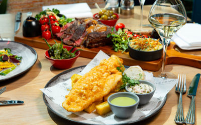 A close-up of a plated meal featuring golden battered fish and thick-cut chips with small bowls of green pea purée and tartar sauce, set on branded paper reading “The Grand Brighton.” In the background, there are other dishes including roasted vegetables, sliced steak, and a glass of white wine on a wooden restaurant table.