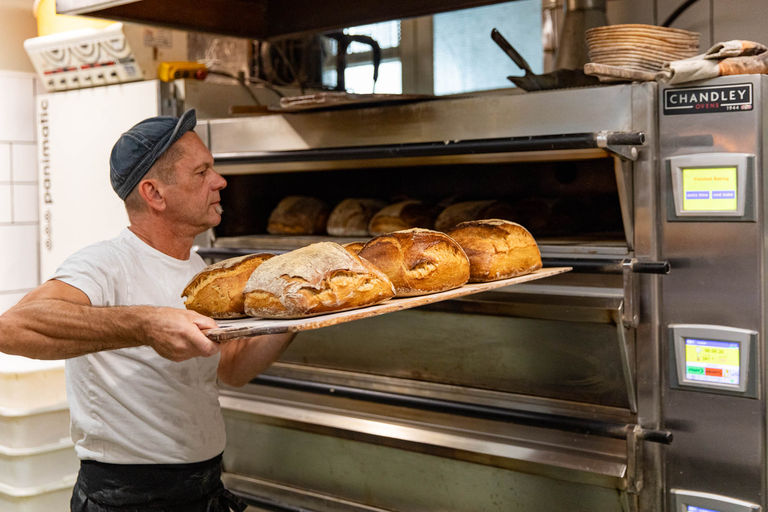 A baker in a white tshirt and a blue hat takes out a batch of cooked loafs from a large industrial oven