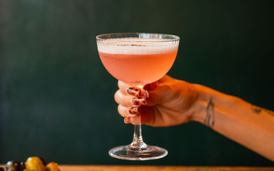 hand holding peach colored cocktail with the dark green background