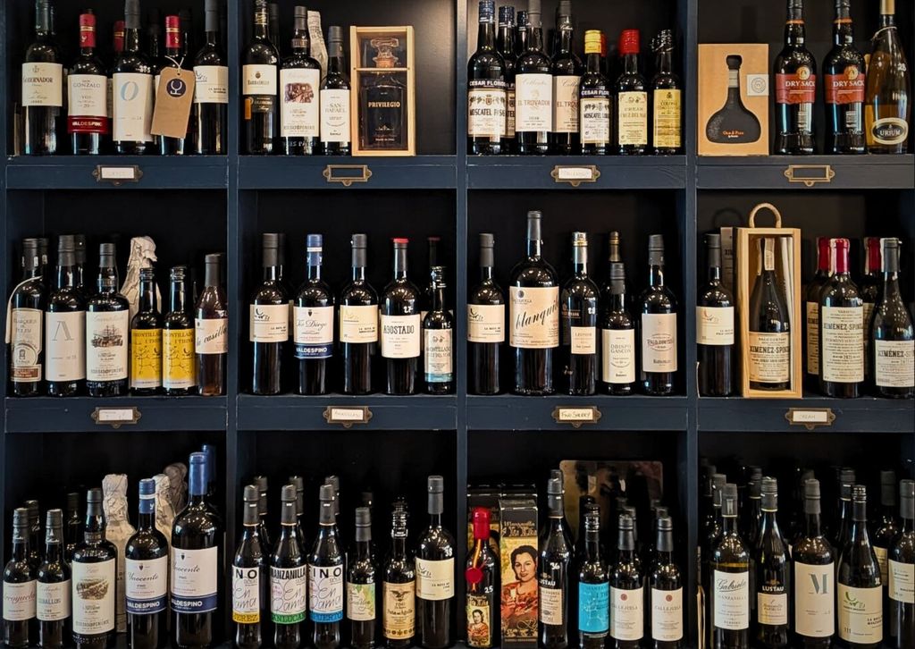 A dark blue shelving unit with 12 alcoves holds many different bottles of sherry. Some are in presentation boxes, others are wrapped. The bottles vary in size and design.