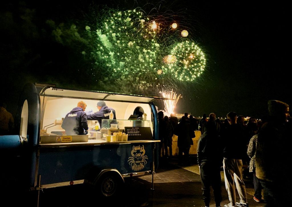 A food trailer is lit up on a dark night as it prepares food for people spectating a fireworks display. You can see the shadows of people as they watch a flurry of green and golden fireworks in the night sky.