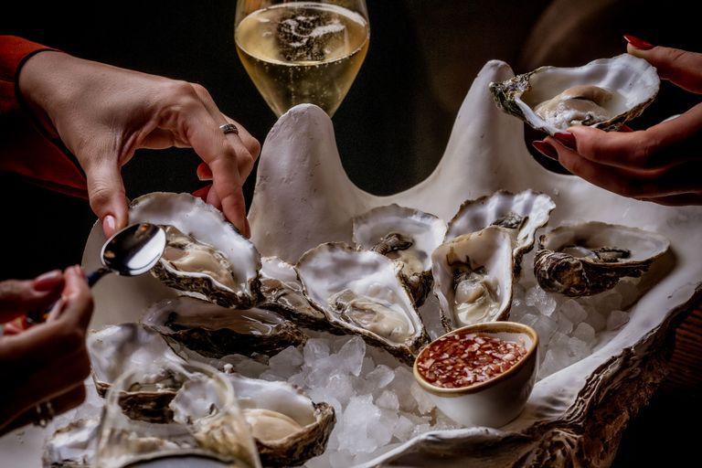A huge oyster shell styled bowl is filled with ice and holds real oysters shucked and ready to eat. Hands hover over to indulge in the feast. There is wine one the table and a vinigerette for the oysters