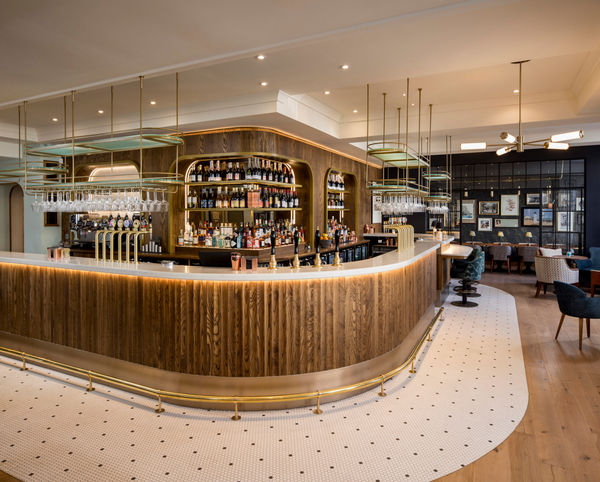 A bright, modern bar interior. In the foreground is a curved, white marble-top bar counter, with a front of warm wood panels and a brass foot rail. Above the bar hang multiple glass racks from brass-and-pale-green metal frames, and glasses are suspended within them. Behind the bar is a well-stocked shelving unit set into a dark wood-panelled wall, filled with bottles of spirits and wine. To the right of the bar counter, seating begins: upholstered chairs and small tables sit on a light wood floor, and a section of the floor in front of the bar is tiled in white hexagon tiles with small dark dots. At the far right, a dark accent wall displays framed pictures and there are lounge-style chairs and booths, generating a relaxed atmosphere. The ceiling is light in colour, recessed with spotlights, and a modern chandelier with brass arms and white shades hangs above the seating area.