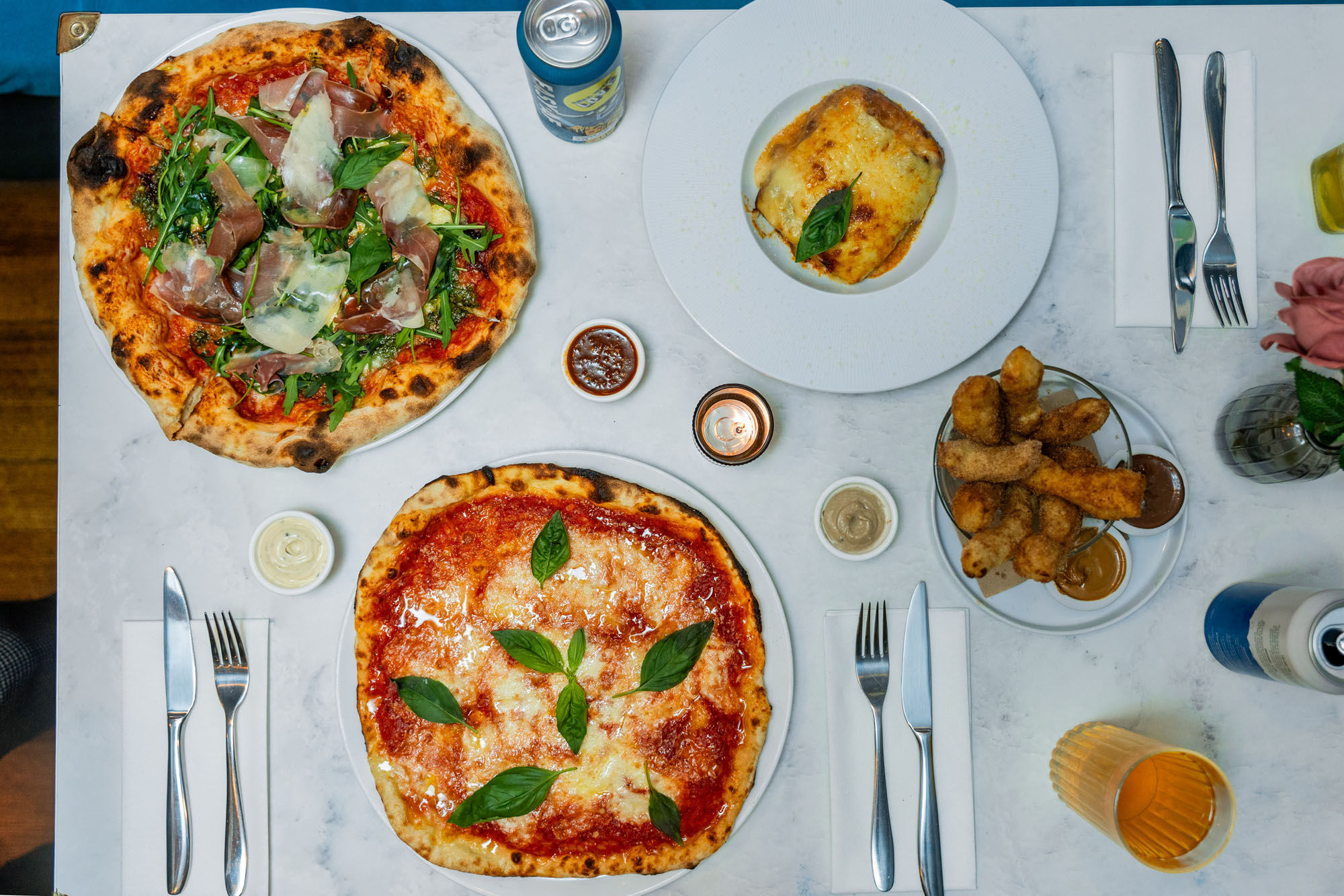 over head shot of the white table laid our with couple of dishes including pizzas, lasagna and drinks