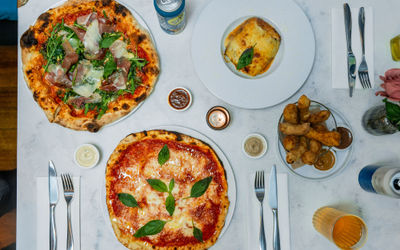 over head shot of the white table laid our with couple of dishes including pizzas, lasagna and drinks