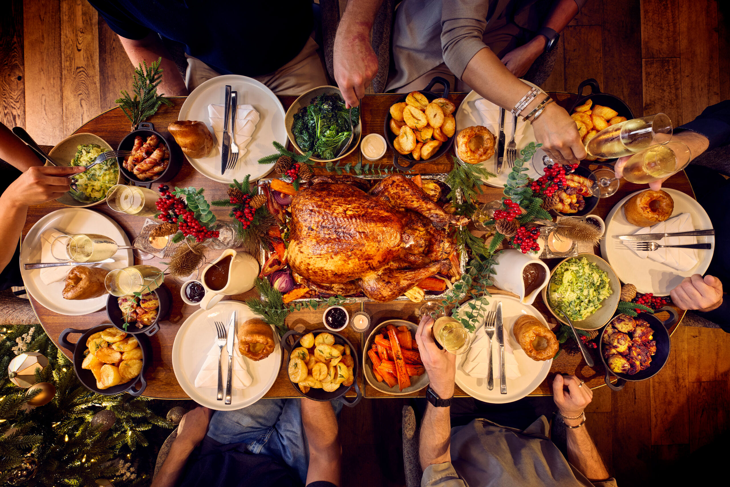 A dining table for 6 is set for christmas. The photography is from above and shows a whole festive spread with a glazed turkey being in the centre of the table. Places are set with white plates and silver cutlery. Surrounding the turkey are bowls of trimmings from brussel sprouts, carrots and roast potatoes. it looks like a proper feast.
