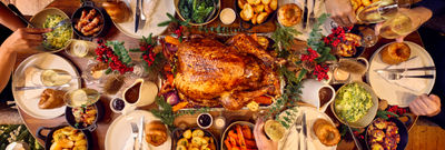 A dining table for 6 is set for christmas. The photography is from above and shows a whole festive spread with a glazed turkey being in the centre of the table. Places are set with white plates and silver cutlery. Surrounding the turkey are bowls of trimmings from brussel sprouts, carrots and roast potatoes. it looks like a proper feast.