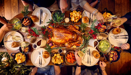 A dining table for 6 is set for christmas. The photography is from above and shows a whole festive spread with a glazed turkey being in the centre of the table. Places are set with white plates and silver cutlery. Surrounding the turkey are bowls of trimmings from brussel sprouts, carrots and roast potatoes. it looks like a proper feast.