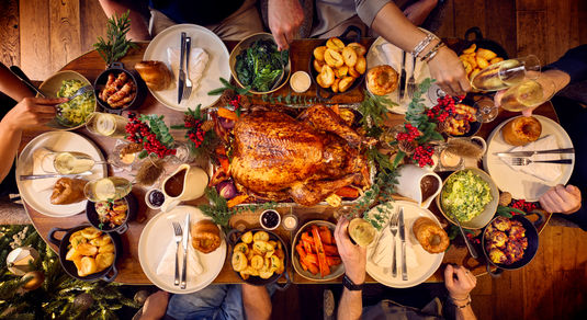 A dining table for 6 is set for christmas. The photography is from above and shows a whole festive spread with a glazed turkey being in the centre of the table. Places are set with white plates and silver cutlery. Surrounding the turkey are bowls of trimmings from brussel sprouts, carrots and roast potatoes. it looks like a proper feast.