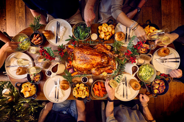 A dining table for 6 is set for christmas. The photography is from above and shows a whole festive spread with a glazed turkey being in the centre of the table. Places are set with white plates and silver cutlery. Surrounding the turkey are bowls of trimmings from brussel sprouts, carrots and roast potatoes. it looks like a proper feast.