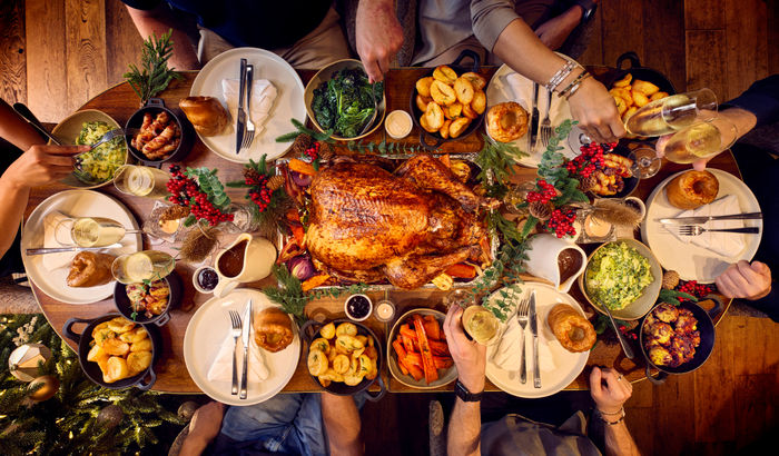 A dining table for 6 is set for christmas. The photography is from above and shows a whole festive spread with a glazed turkey being in the centre of the table. Places are set with white plates and silver cutlery. Surrounding the turkey are bowls of trimmings from brussel sprouts, carrots and roast potatoes. it looks like a proper feast.