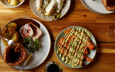 A wooden table holds several plates of sunday roast food. From whole carrots drizzled in sauce to a vuts of meat with gravy. There is a glass of red wine in the mix. It looks inviting and appetising.